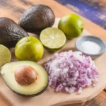 A close-up of fresh avocados, limes, chopped onions, and salt for making guacamole.